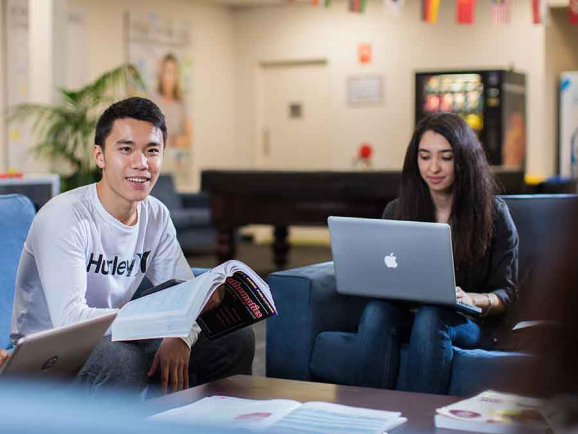 Male student reading a text book, female student on laptop, sitting in common area couches