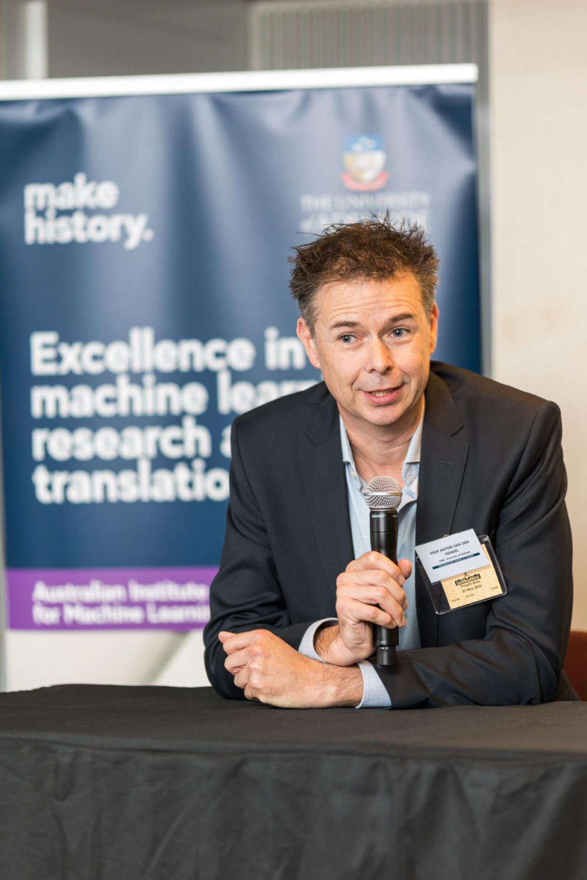 anton van den hengel speaking with microphone at parliament house event with university of adelaide poster behind