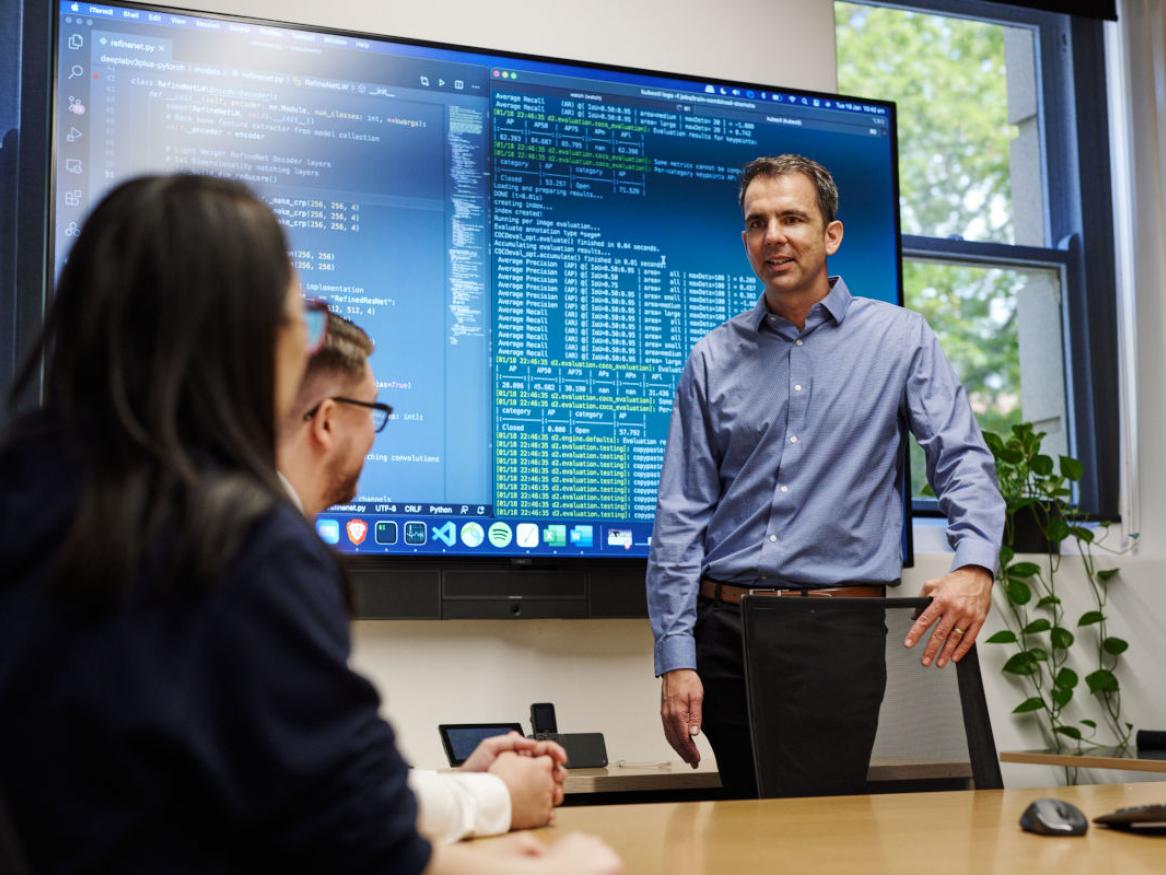 AIML's director Professor Simon Lucey standing next to a large LED screen with computer code on it