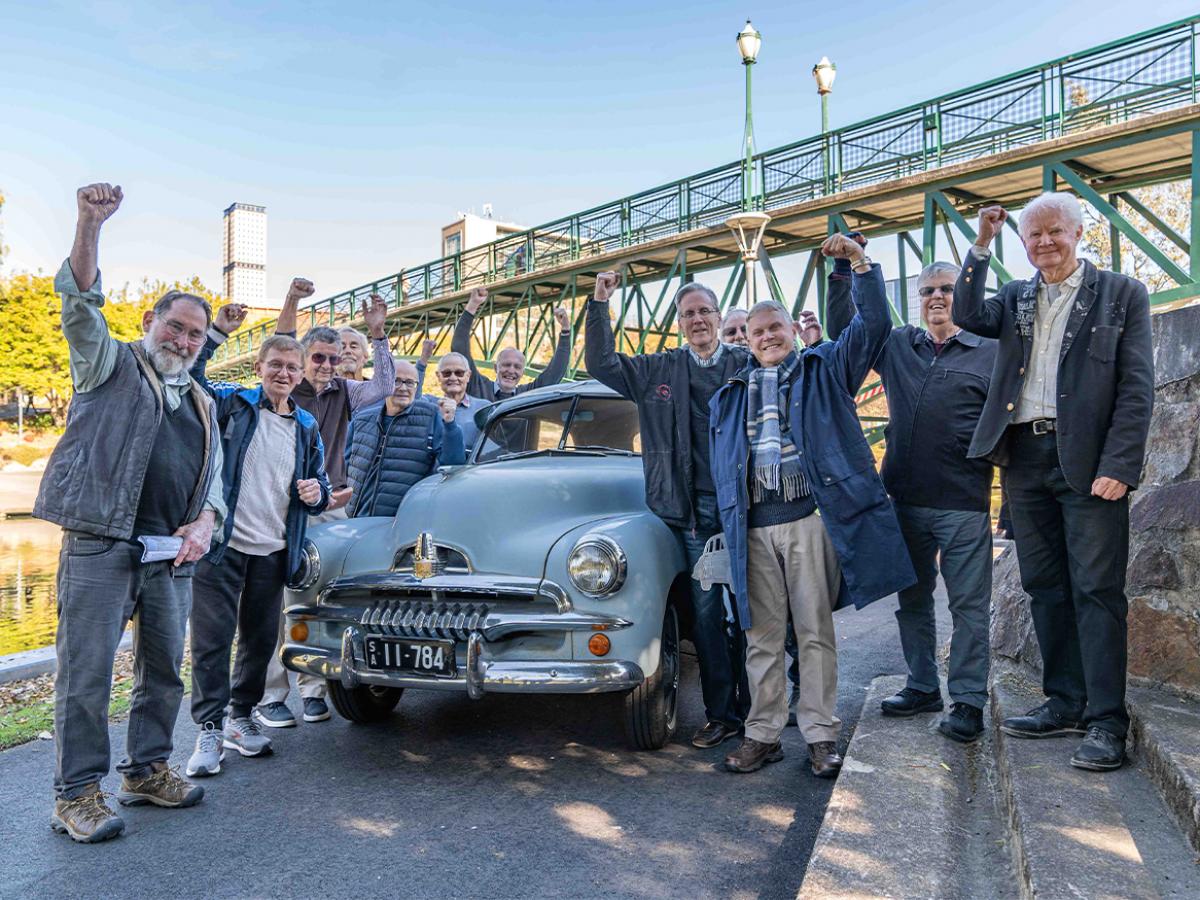 12 engineering alumni around an old dusty blue FJ car at the uni footbridge
