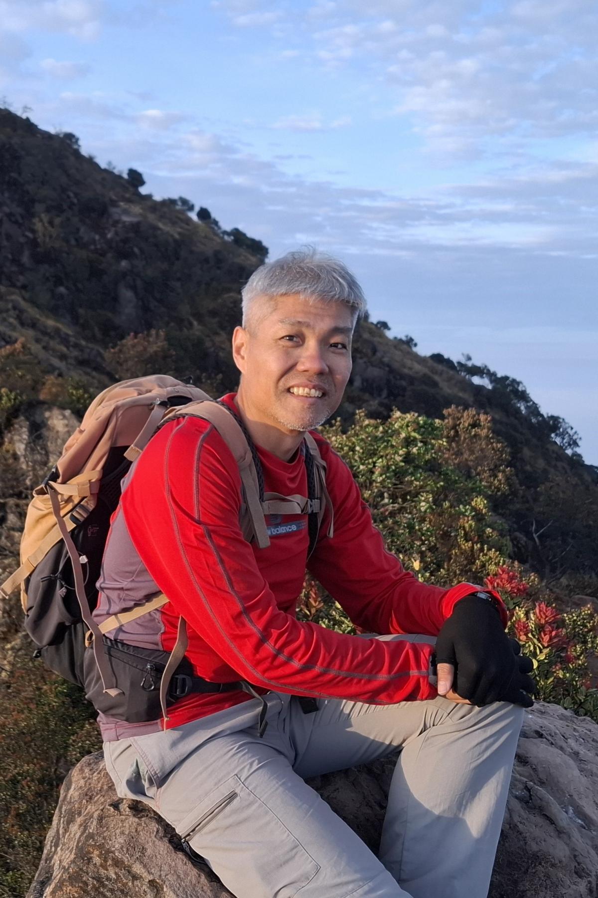 A portrait of Sam Ng sitting on a rock, on a mountain