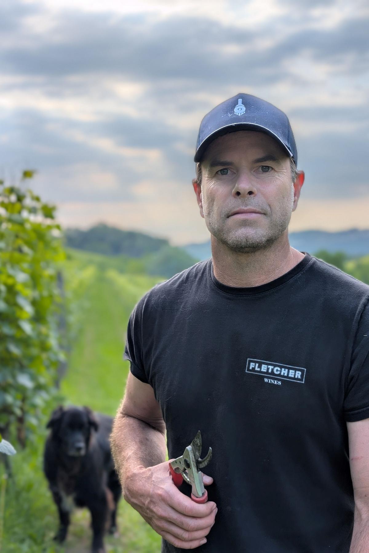 Dave is in a vineyard, wearing a hat, holding a pair of secateurs, with a dog in the background
