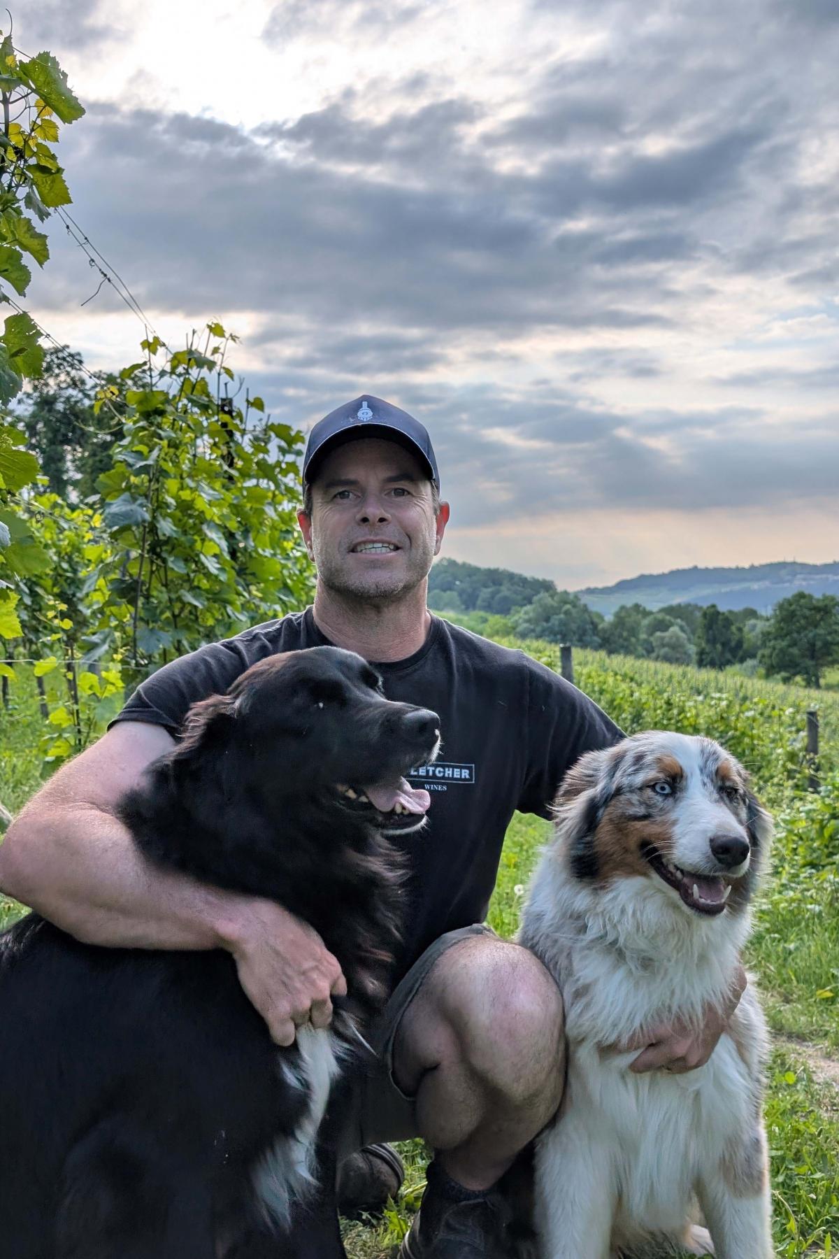 A photo of Dave Fletcher smiling with two large dogs in a vineyard