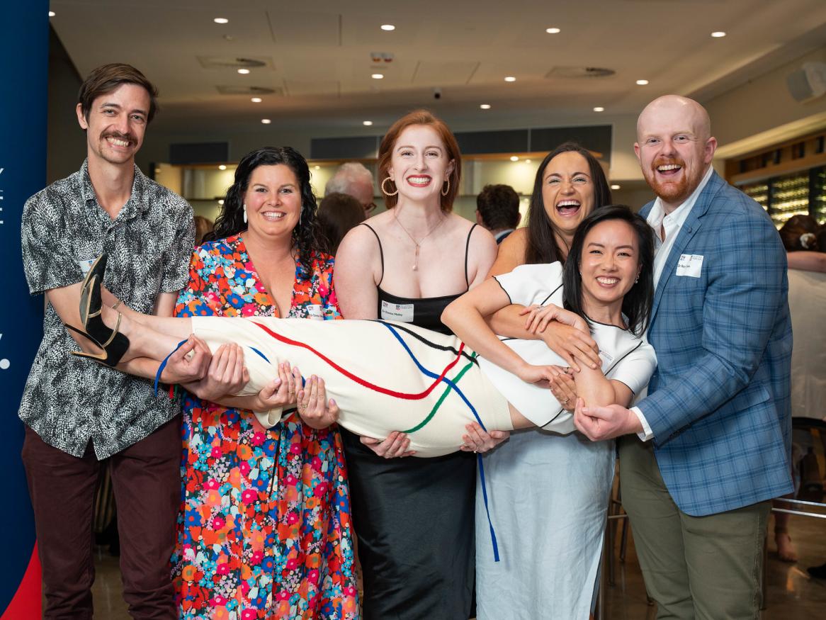 Five laughing committee members hold up a sixth members 