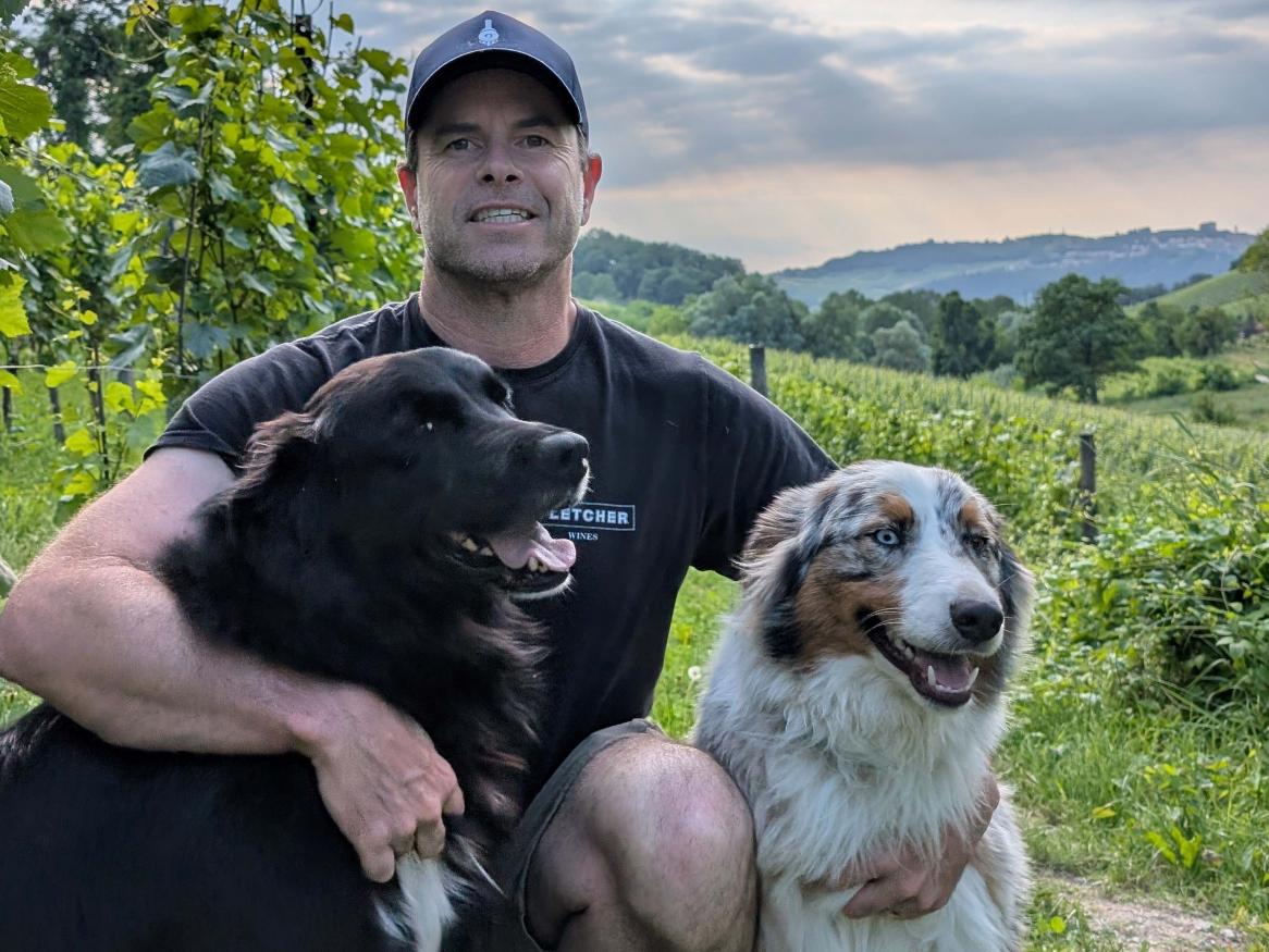 A photo of Dave Fletcher smiling with two large dogs in a vineyard