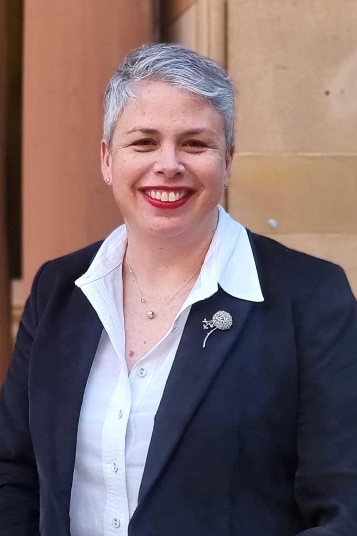 Lori is wearing a dark blazer over a white shirt, and is smiling into the camera