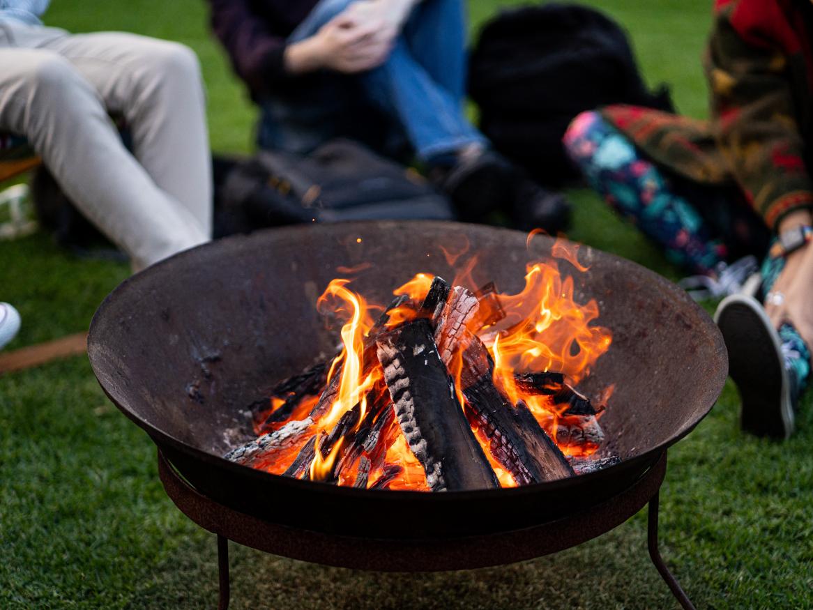 A fire burns in a fire pit, with University community members sitting around it