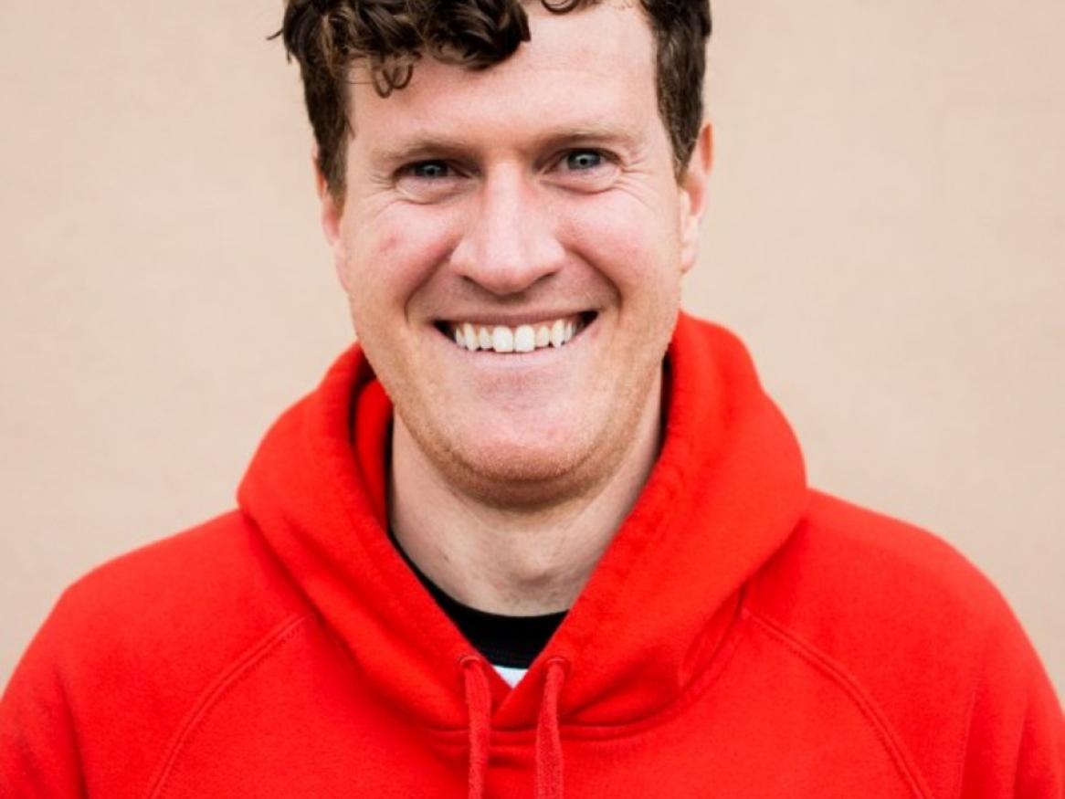 A young man wearing a bright red hoodie jumper is smiling at the camera