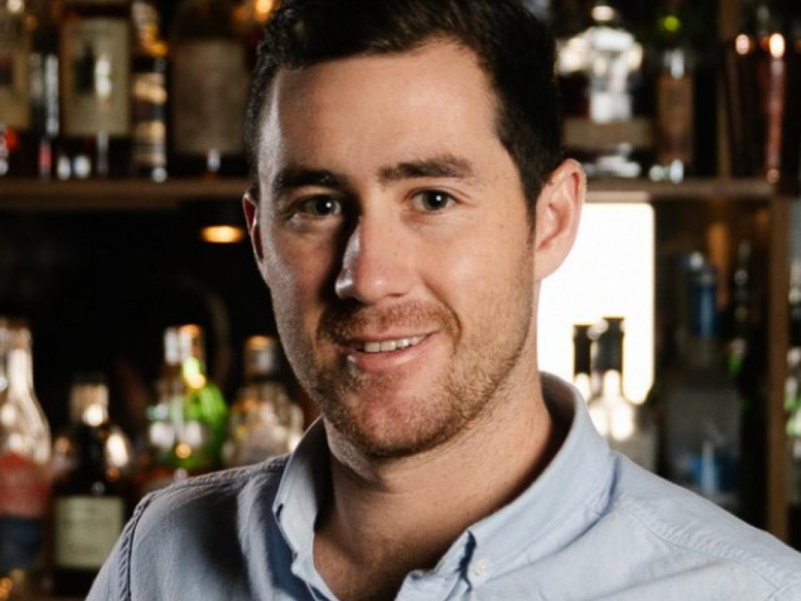 A young man stands in front of  a bar with bottles of alcohol in the background, smiling at the camera