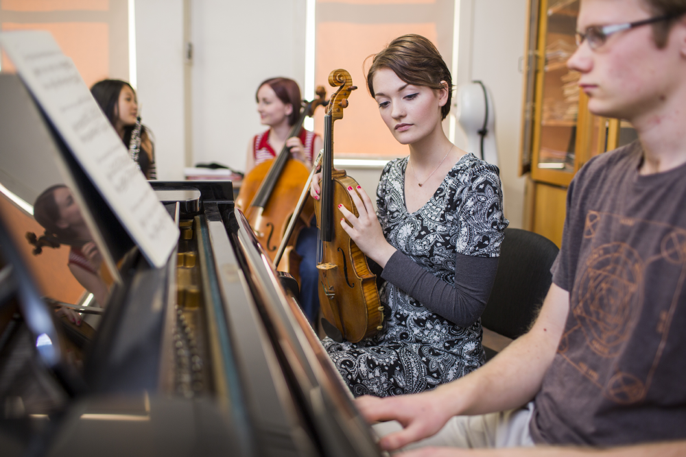 Music students sitting together, one at a piano, one holding a violin and one holding a cello