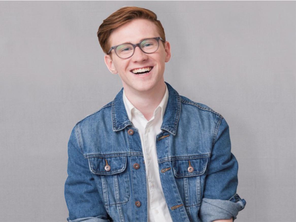 Young male student is smiling at the camera, wearing glasses and a denim jacket.