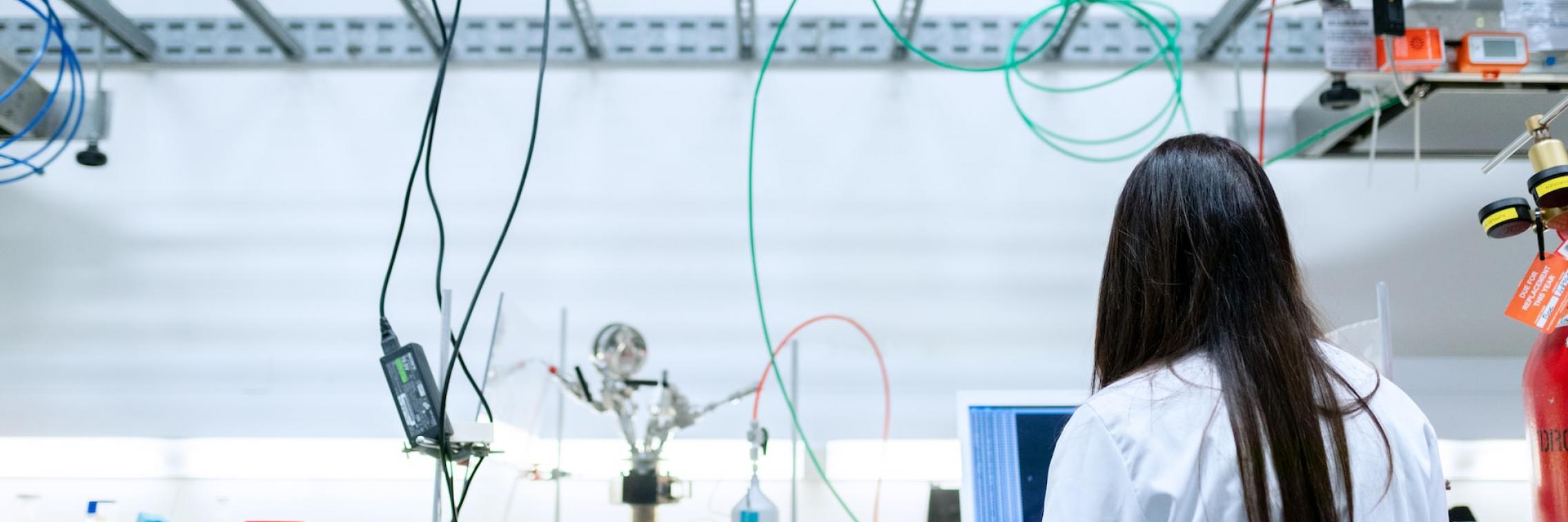 A woman with her back to the camera conducts scientific experiments; on the bench in front of her stand numerous scientific instruments.