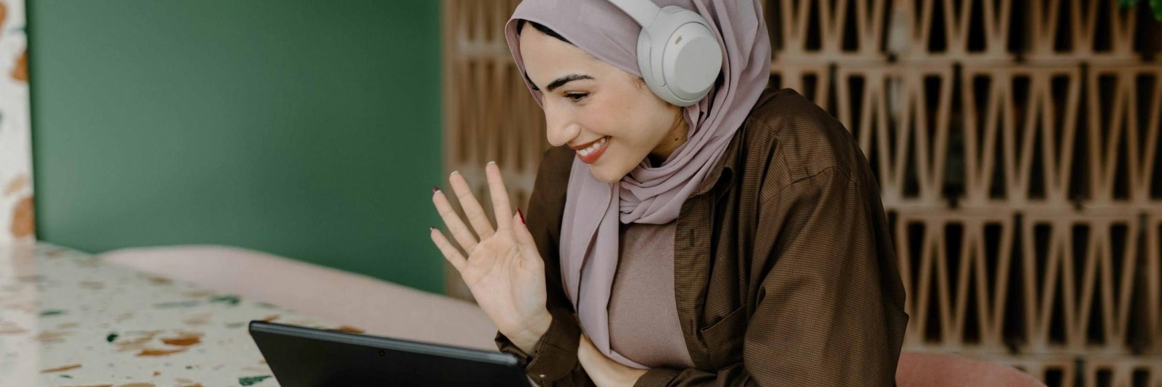 A woman with headphones waves at the camera on their laptop computer.