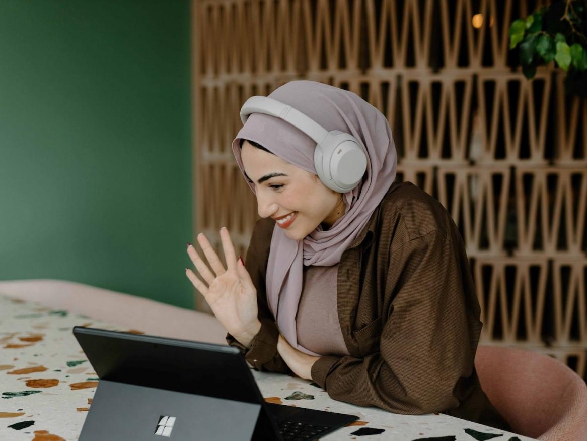 A woman with headphones waves at the camera on their laptop computer.