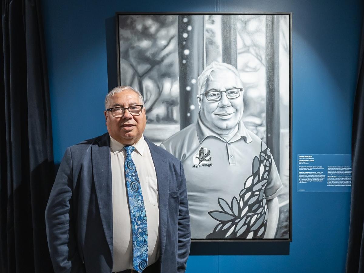 Uncle Rod stands with his portrait: Uncle Rodney O’Brien, Kaurna Elder (2021) by Thomas Readett, Ngarrindjeri/Arrernte people. University of Adelaide Library (A.VA.2022.1031)