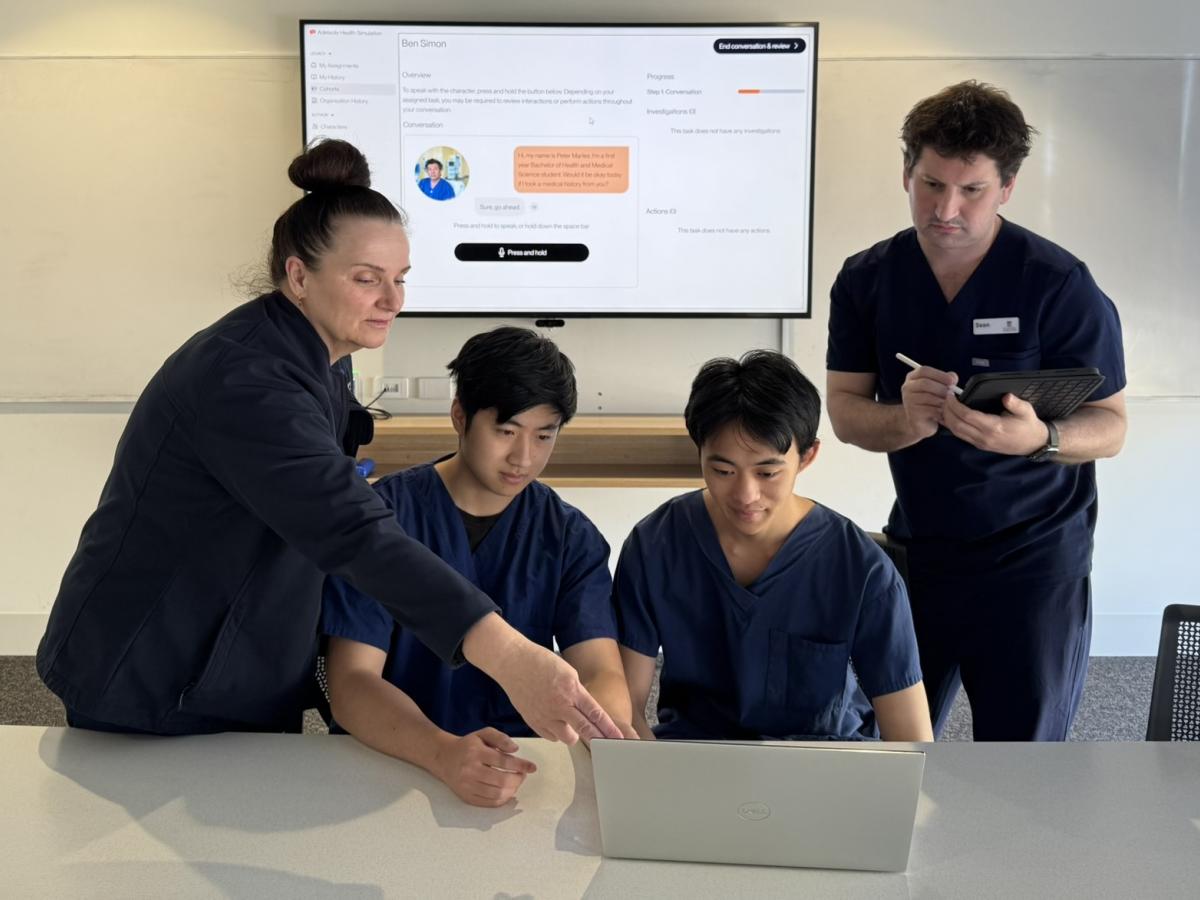 Four people in a classroom; two students in scrubs at a laptop, guided by two educators.