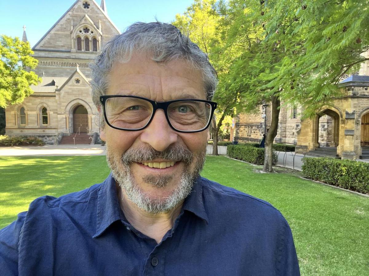 A man with glasses and a blue shirt smiles at the camera while standing on a lawn in front of the historic stone Elder Hall building at the University of Adelaide.