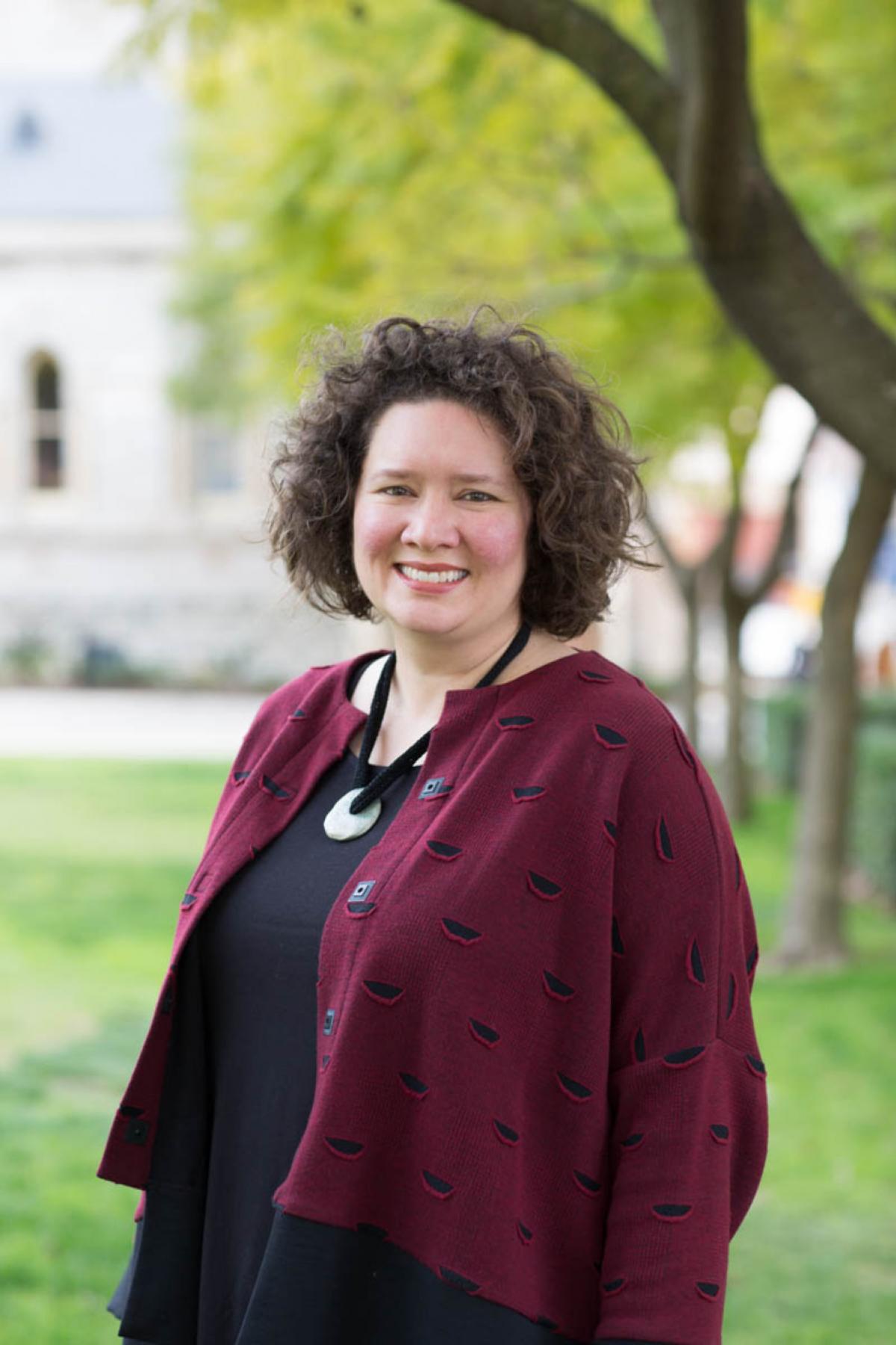 A woman with curly hair smiling, wearing a maroon cardigan and necklace, standing outdoors with trees and grass in the background.