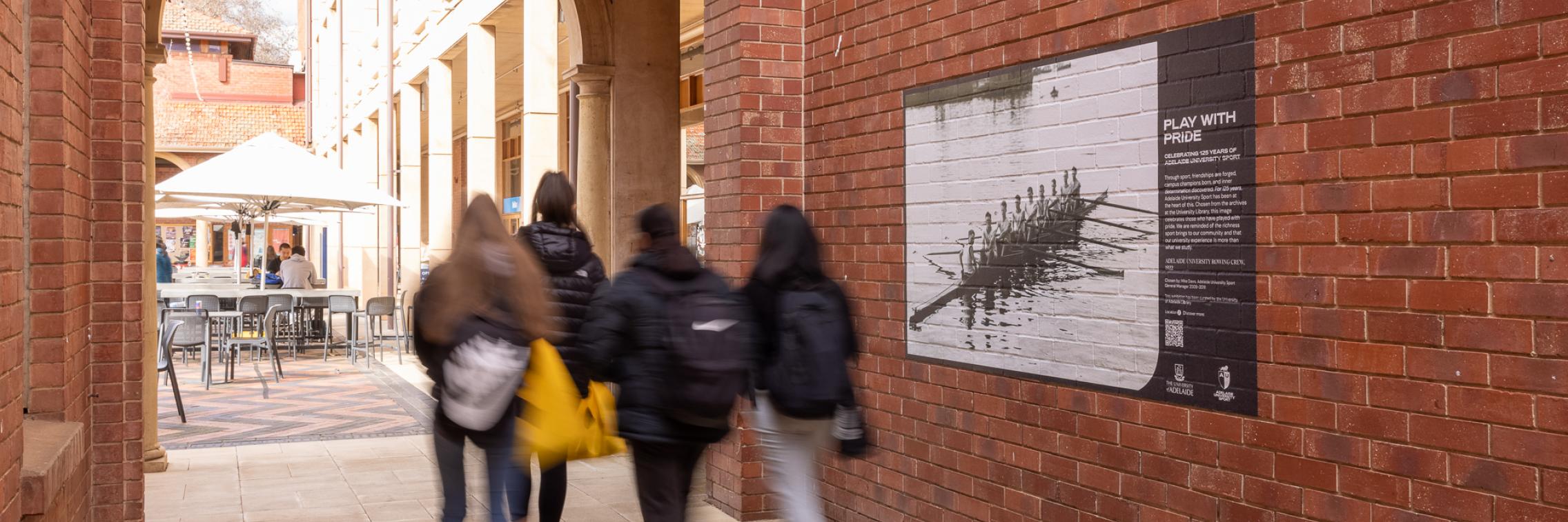 Students walking past Play with Pride Exhibition image pasted up on brick wall