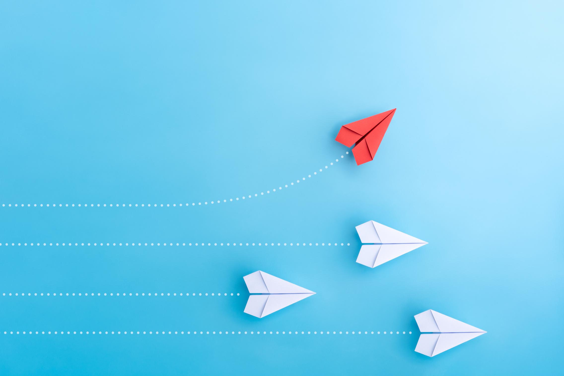Photo of a red paper plane flying away from a group of white paper planes.