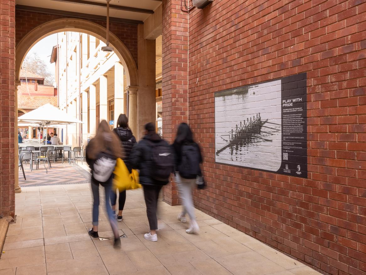 Students walking past Play with Pride Exhibition image pasted up on brick wall