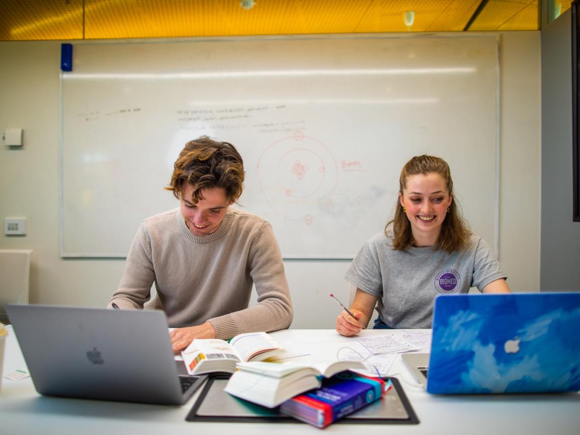 Two students sitting in front of a whiteboard with their laptops and study papers