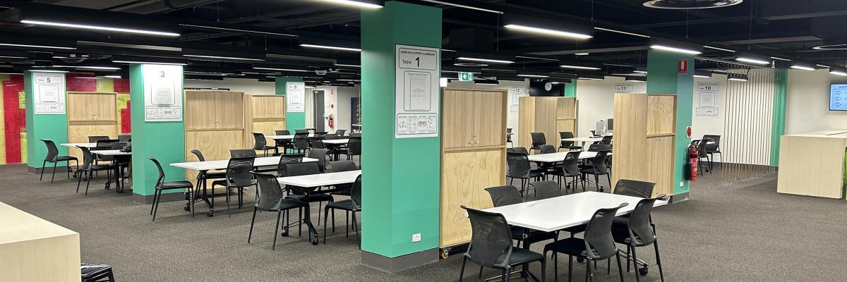 A room with white tables each with six black chairs. Supporting pillars are painted green and each have a poster declaring a table number. There are also wooden cupboards dividing the space into smaller classrooms.