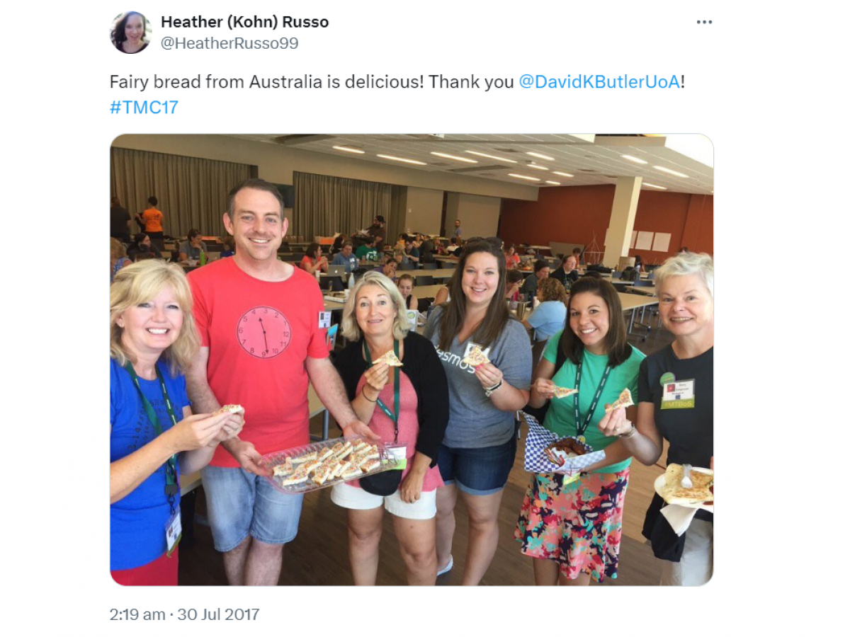 A Twitter post from Heather (Kohn) Russo @HeatherRusso99 on 30 Jul 2017 with text and a photo. The text says: Fairy bread from Australia is delicious! Thank you @DavidKButlerUoa #TMC17 The photo contains: Six people smiling at the camera. Behind us is a big room with chairs and tables and lots of other people. I am the second person from the left, holding a tray of fairy bread. The other people are all holding a triangular piece of fairy bread.