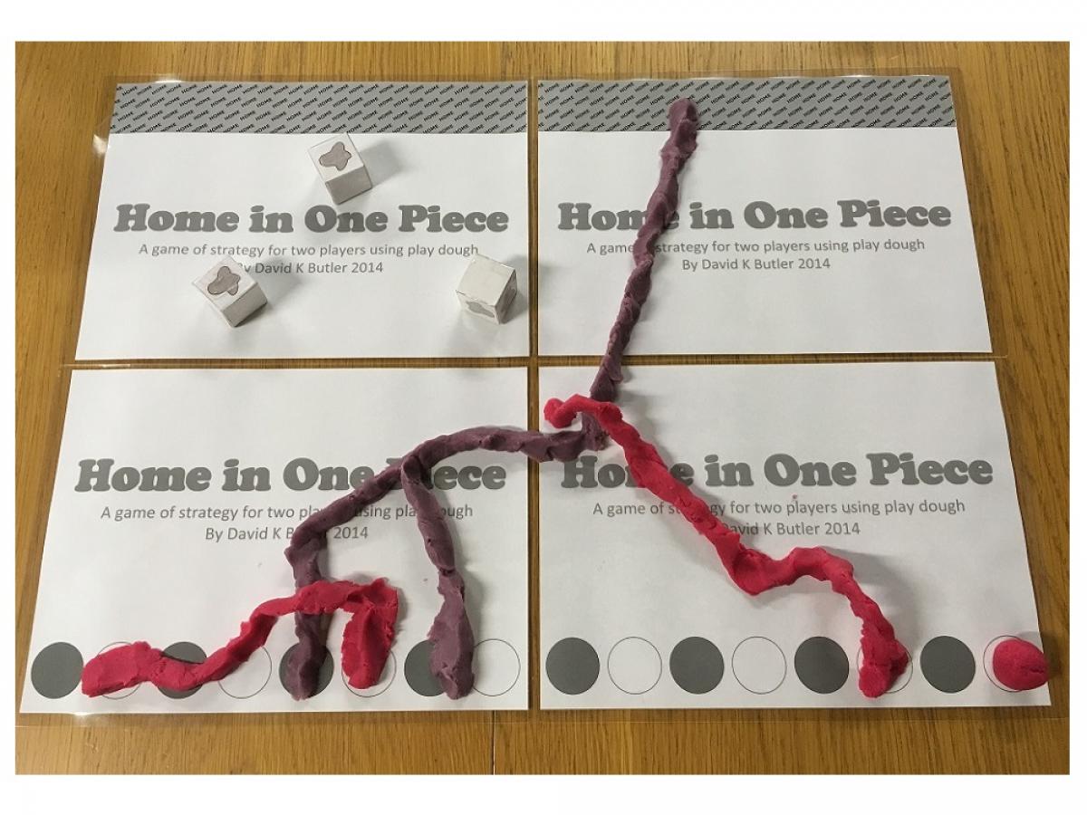 An image of the Home in One Piece board. At the top is a grey zone, at the bottom are white and grey circles alternating. The board has wierdly shaped balls and tendrils of pink and purple play dough over it. You can also see three dice with blob shapes on the faces.
