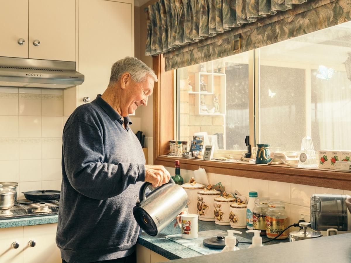 An older man smiles in the kitchen as he makes a cup of tea.