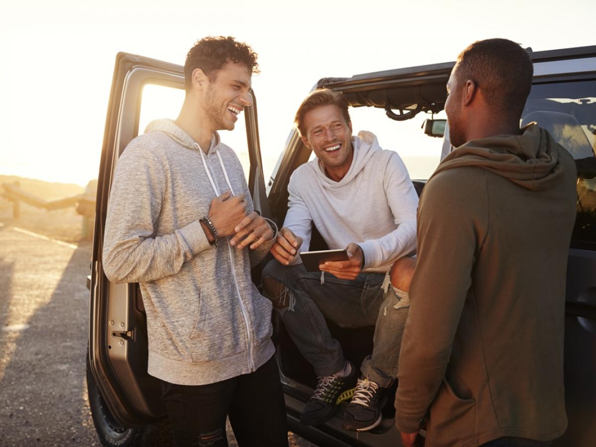 Stock image of three men. One is seated in the front passenger side of the car with the door open holding a tablet. the other two are standing either side of him.
