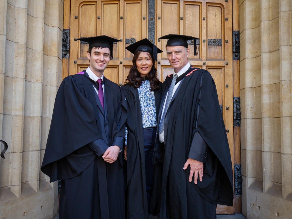 David, Chharida, and Guy are wearing graduation robes and smiling.