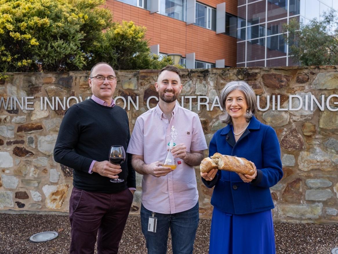 Professor Vladimir Jiranek, Scott Oliphant, and Nicola Spurrier stand holding bread, wine, and bacteria.