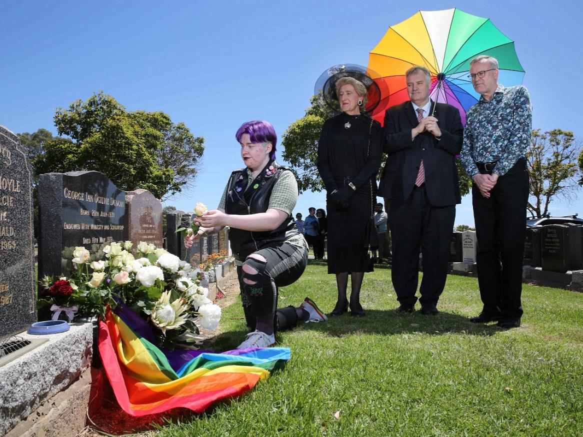 Visitors at Dr Duncan's grave, with a rainbow umbrella, flag, and flowers.