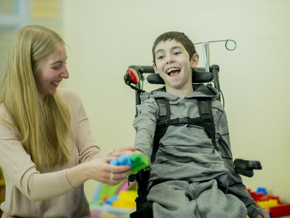 A boy with Cerebral Palsy sits in a wheelchair, smiling and playing with a toy with his carer.