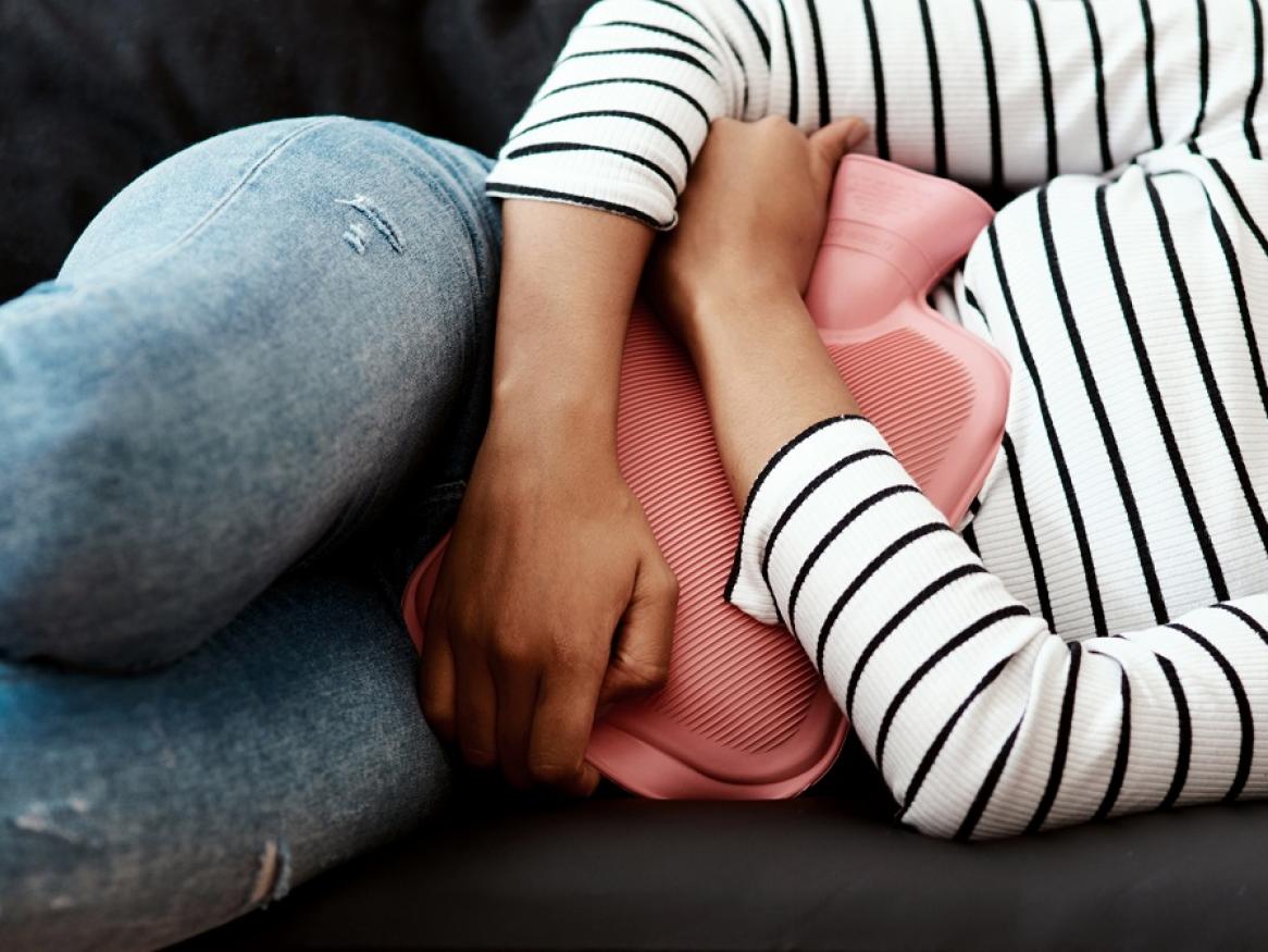 Photo of a women in a white and black stripe top holding a red hot water bottle over her abdomen while lying on a grey couch.