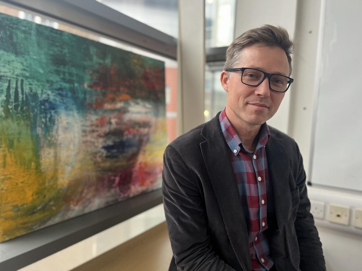 Photo of Professor David MacIntyre. He is wearing a dark blazer and check shirt. He has glasses with dark rims. He is smiling and standing in front of an abstract paiting.