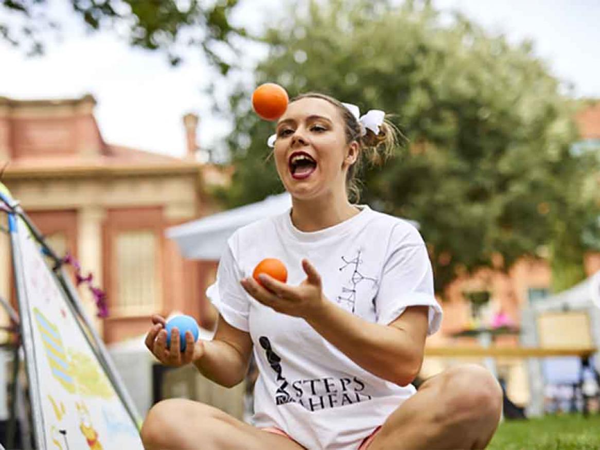 Student sitting on lawn, juggling three balls. 