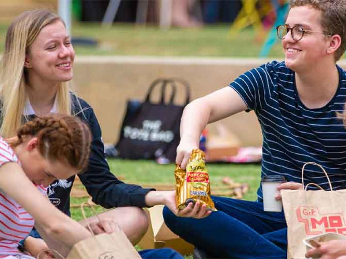 Group of students sitting on campus lawn, eating food, smiling 