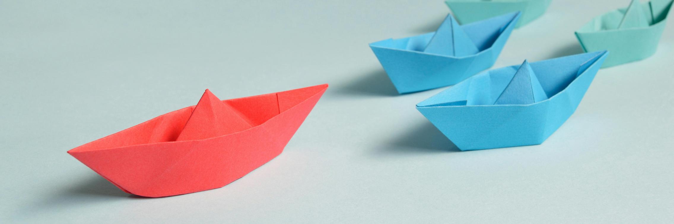 Blue paper boats against a plain background, with a red boat in the lead.
