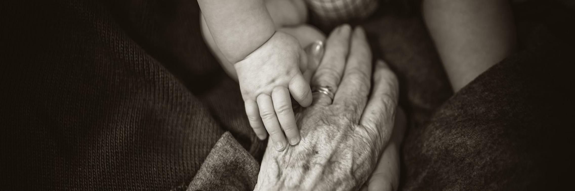 An elderly person holding hands with an infant.