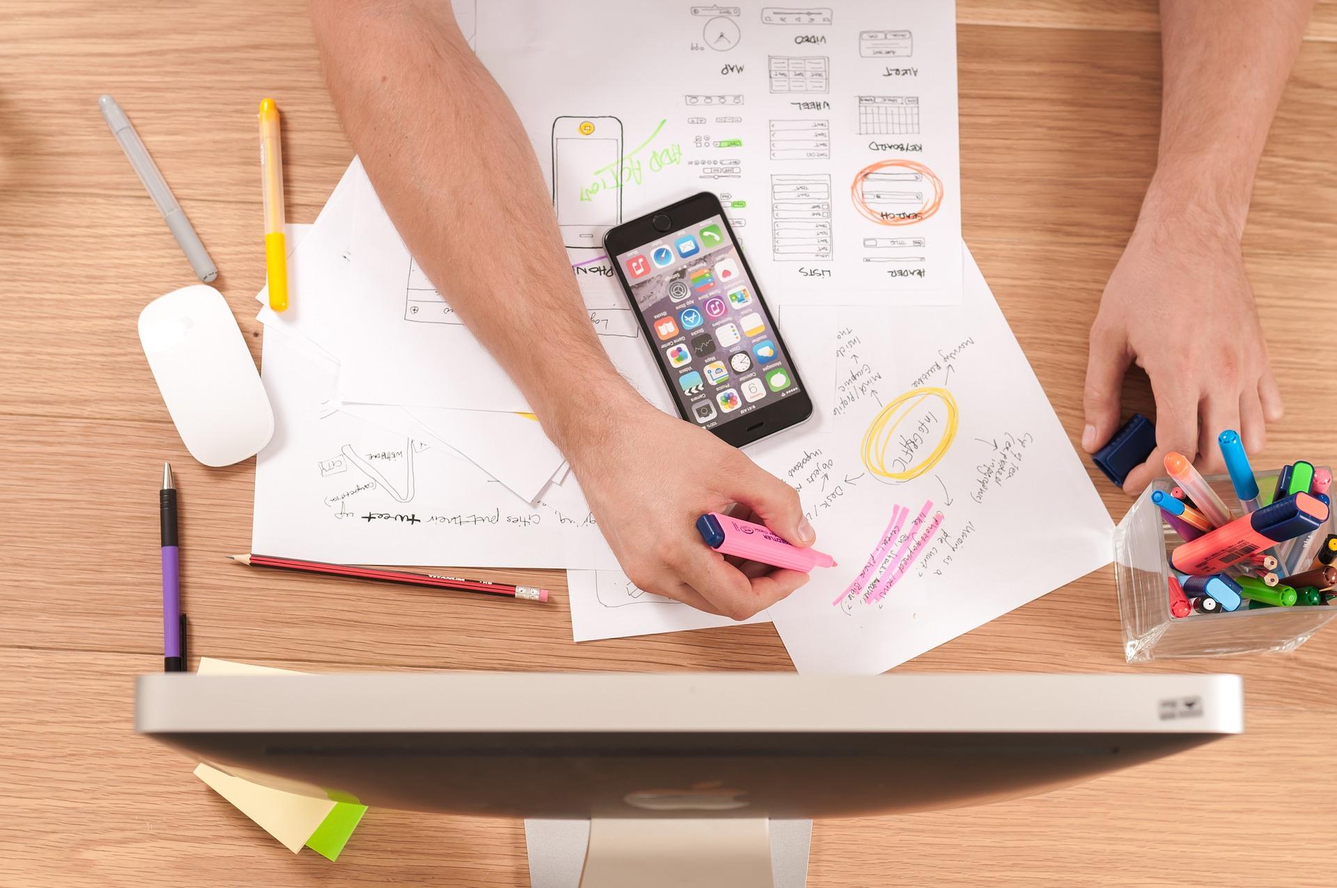 Desk with loose papers and highlighter pens. Someone sits at the desk highlighting a page.