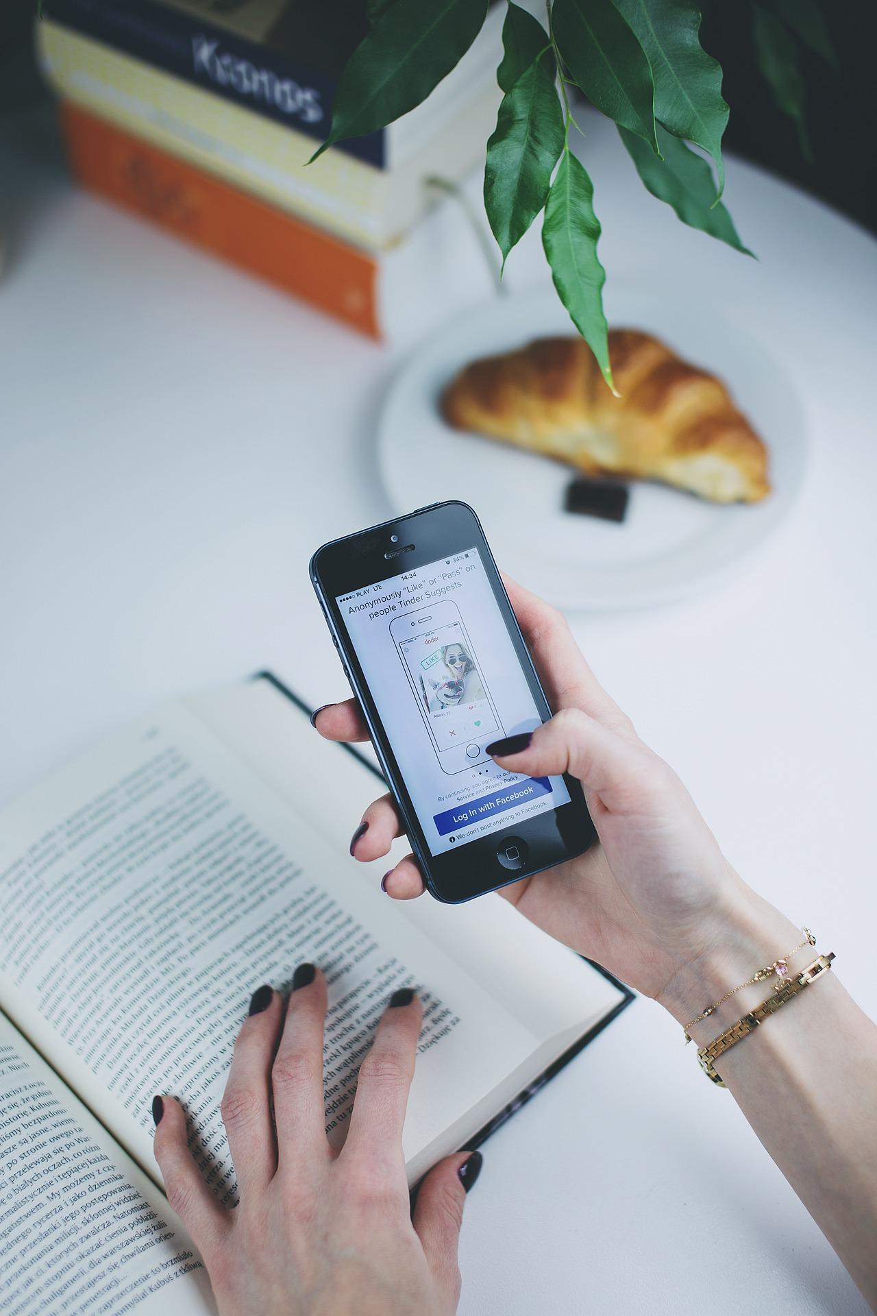 two hands: one resting on a book, the other holding a phone with a dating app