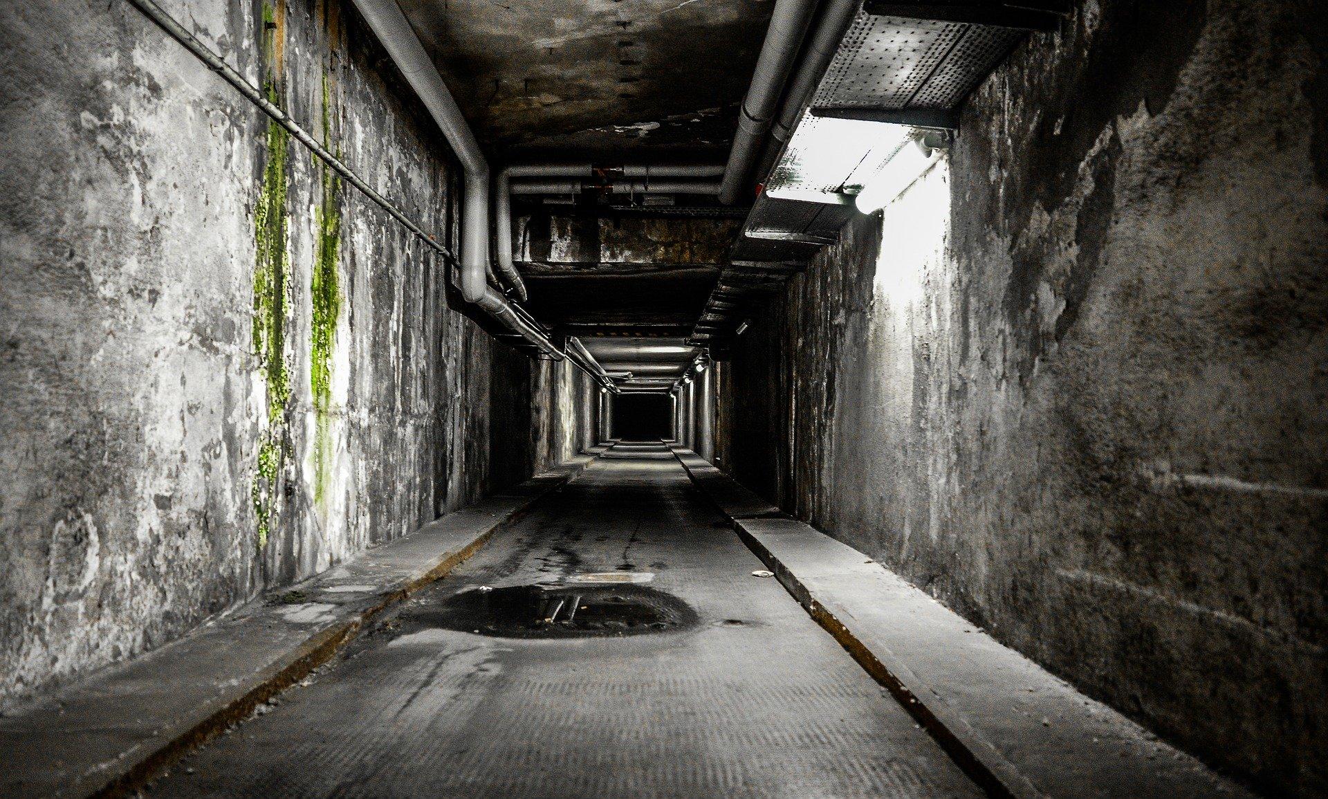 A grey hallway/tunnel with pipes and mold and small flickers of sunlight.