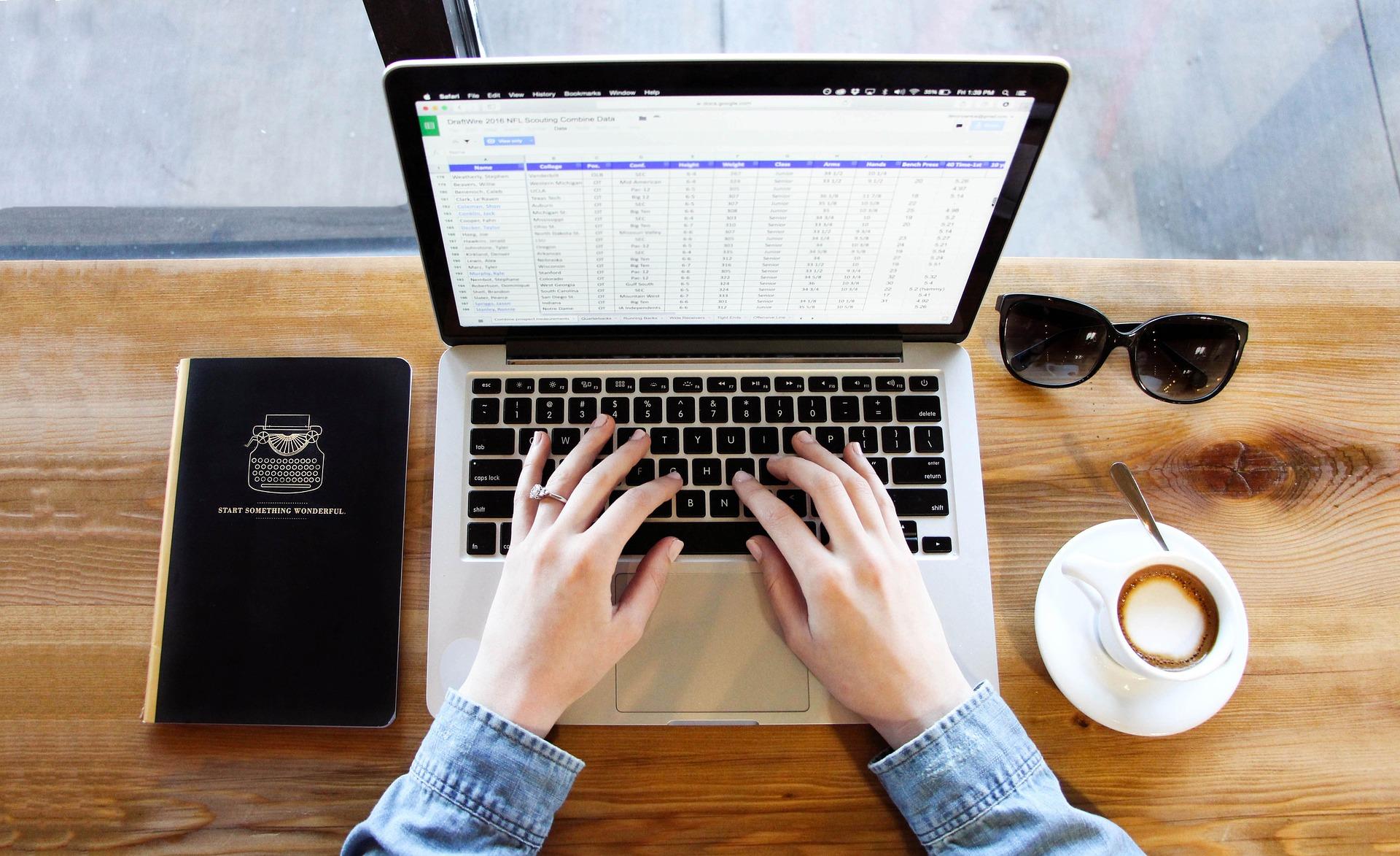 A laptop, coffee cup, sunglasses and notebook on a wooden table with hands hovering over the keyboard