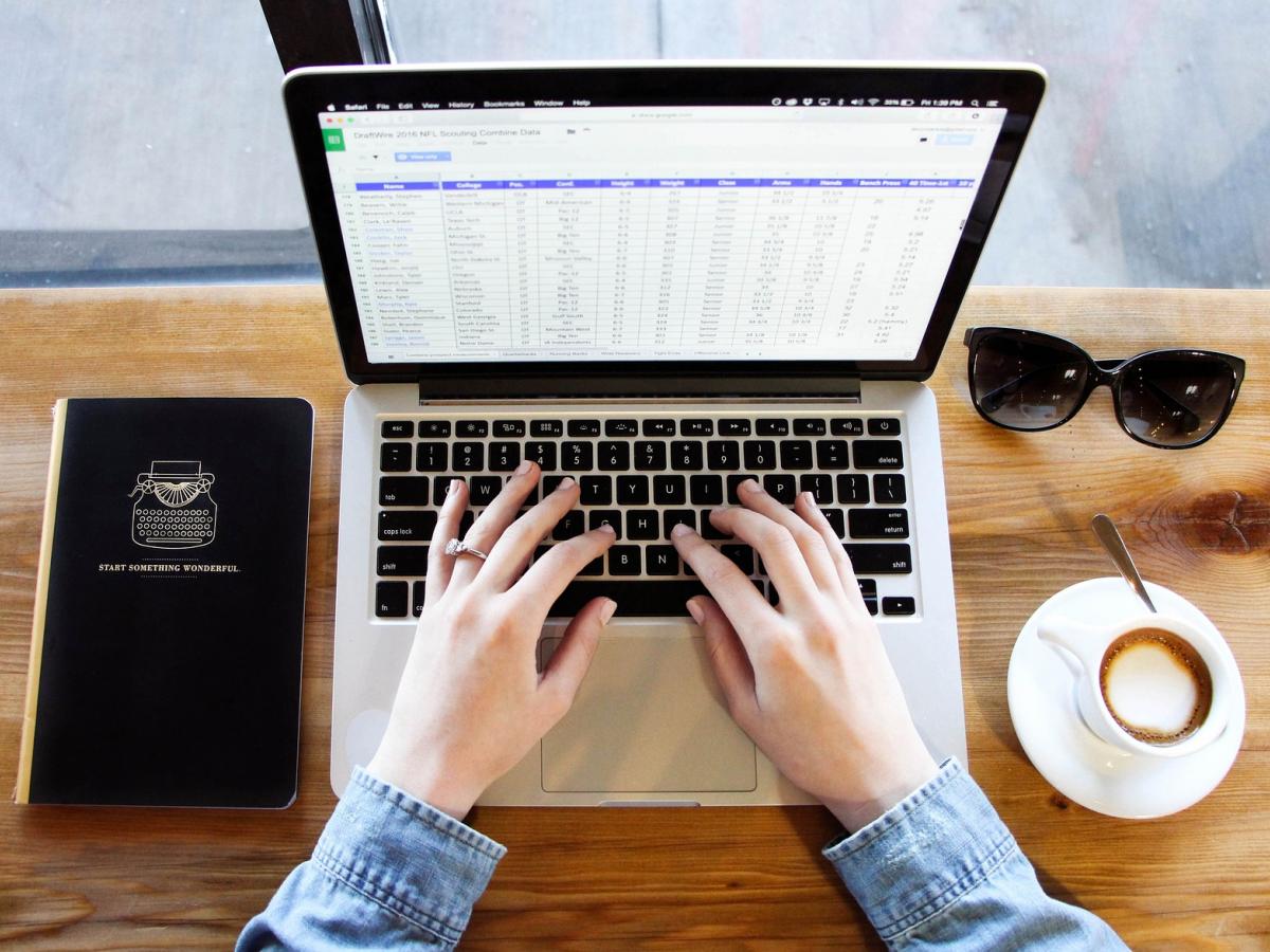 A laptop, coffee cup, sunglasses and notebook on a wooden table with hands hovering over the keyboard