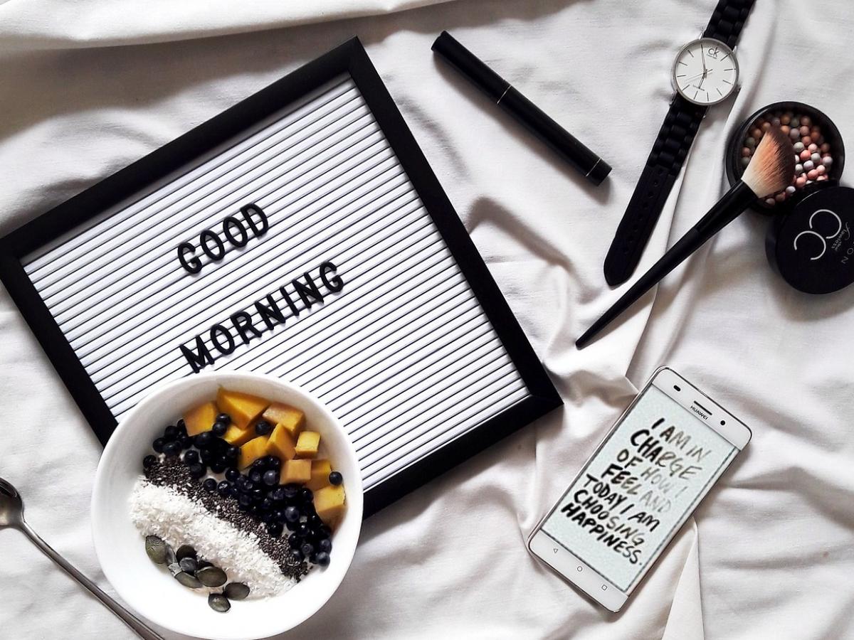 An image of a sheet with a sign reading 'GOOD MORNING', a bowl of fruit, a watch, a phone, and makeup brushes.