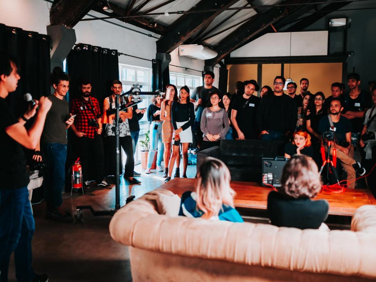 Photo of large group of people at an indoor event, most standing and some sitting on a couch