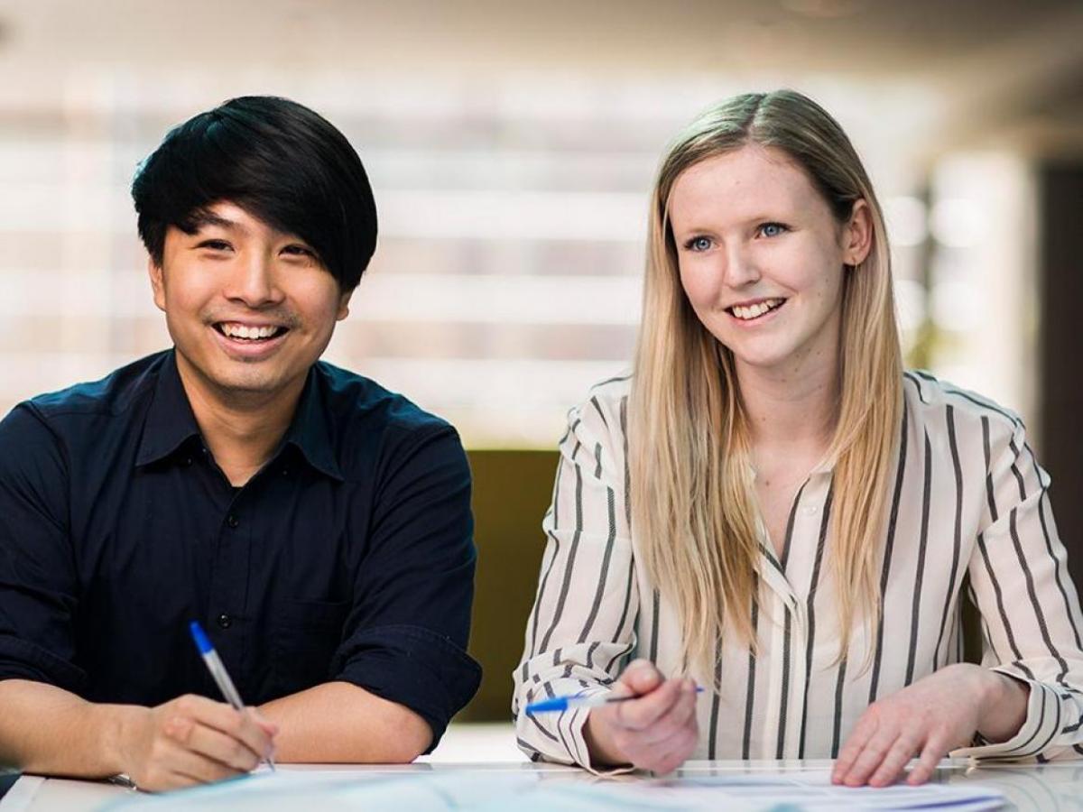 2 people smiling at the camera, with pens and papers on desk