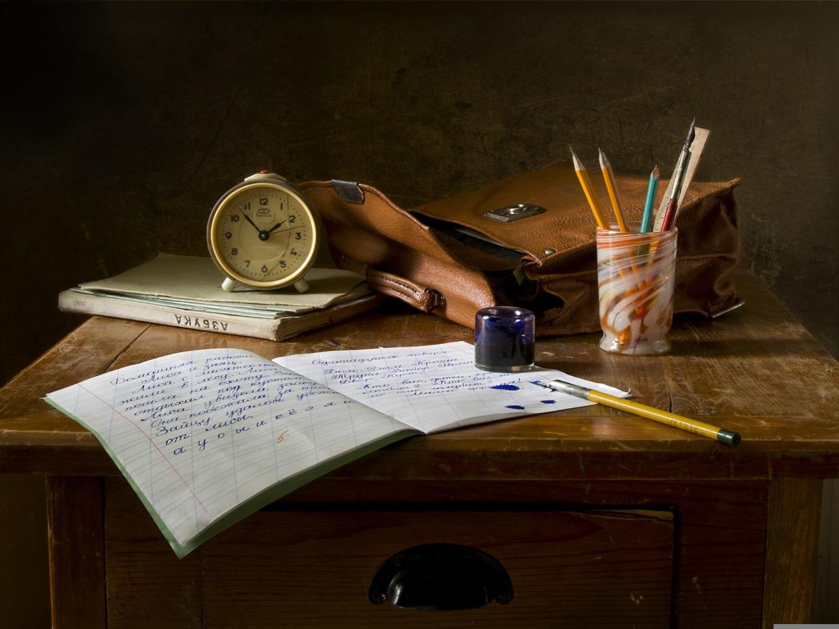 A messy wooden tabletop with some books, an analogue clock, a brown messenger bag, a pot of pencils, as well as ink and a fountain pen.